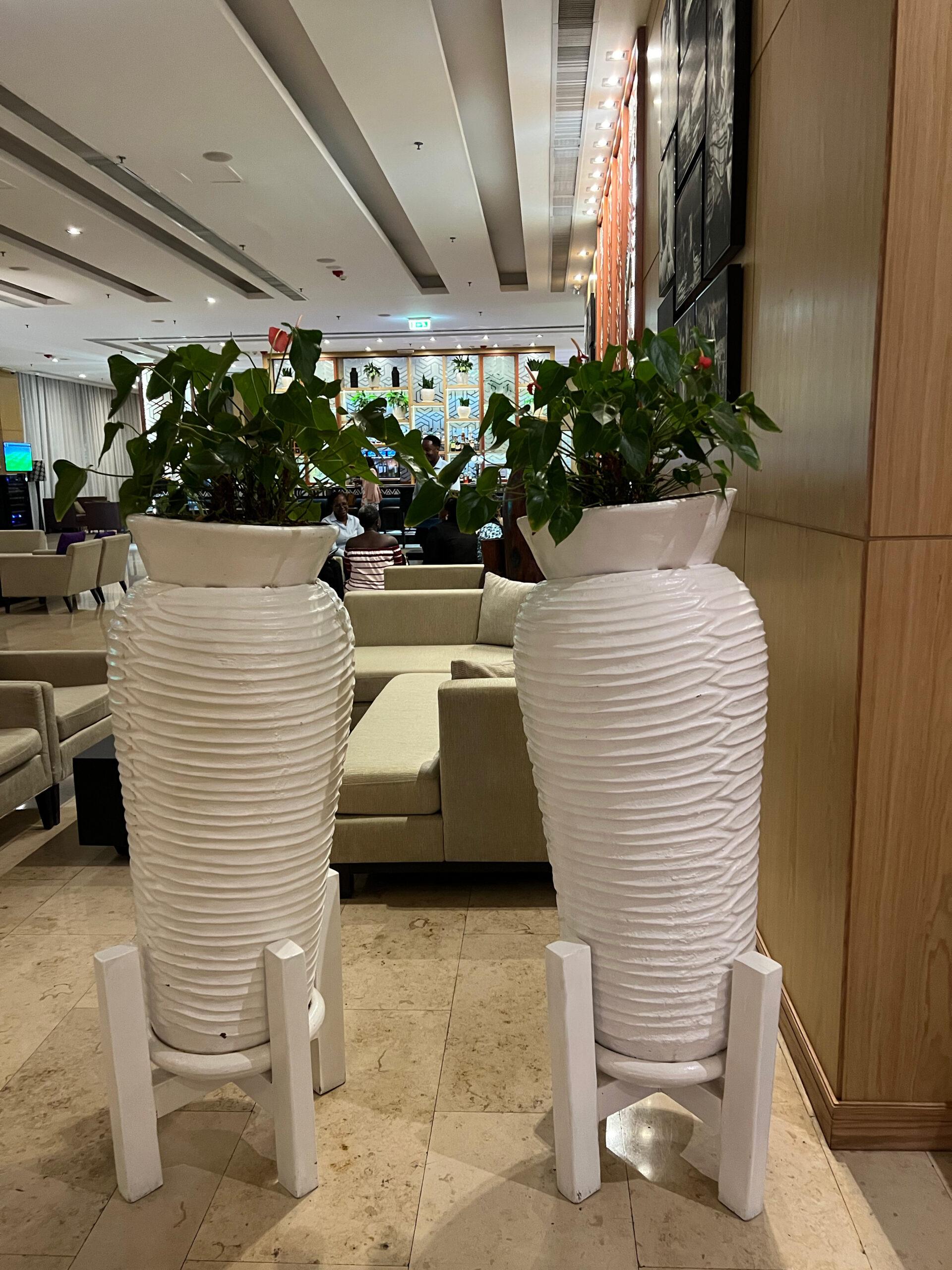 Two large handcrafted African pottery vases with horizontal ribbed texture on wooden cross-base stands, filled with lush green plants, displayed in an elegant interior
